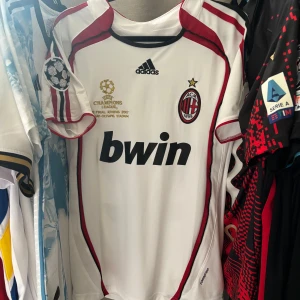 AC Milan Kaka Champions League fotbollströja 2006/07 - AC Milan bortatröja från Champions League-finalen 2007 med Kaka och nummer 22 på ryggen. Vit med röda och svarta detaljer, Adidas-logga, klubbmärke och Bwin sponsortryck. Broderade Champions League-märken på ärmarna. Tillverkad i lätt och ventilerande material.