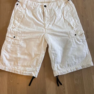 Stone Island shorts  - Snygga vita cargo shorts från Stone Island med flera fickor och svarta detaljer som snörning vid benslut. Tillverkade i bomull för en skön känsla och avslappnad passform. Perfekta för sommardagar när du vill ha en clean och stilren look.