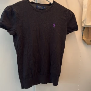 Svart topp från Polo Ralph Lauren - Svart kortärmad topp från Polo Ralph Lauren med rund halsringning och lila broderad logga på bröstet. T-shirten har en klassisk, enkel design och är gjord i mjuk bomull. Perfekt för en stilren och avslappnad look.