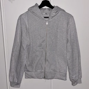 Ljusgrå hoodie med dragkedja - Enkel och stilren ljusgrå hoodie med dragkedja framtill och huva. Tröjan har två fickor på magen och ribbade muddar vid ärmslut och nederkant.