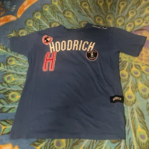 Blå t-shirt från Hoodrich med tryck - Blå t-shirt från Hoodrich med coola tryck och patches framtill. Stor vit HOODRICH-text, röd H och svarta märkespatches ger streetkänsla. Klassisk passform .