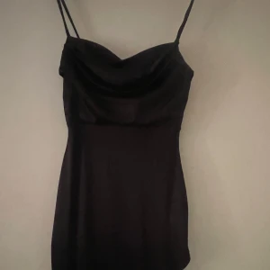 Svart långklänning med tunna axelband - Elegant svart långklänning med tunna axelband lår-hög slits och draperad urringning. Klänningen har en figurnära passform och är tillverkad i ett glansigt, slätt stretchig satin material som ger en lyxig känsla. Nypris:900. Storlek s materialet är ned till marken även med klackar. 