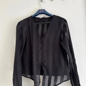 Svart transparent randig skjorta med knyt - Snygg svart skjorta från H&M i transparent material med breda ränder. Skjortan har v-ringning, knappar framtill och långa ärmar. 