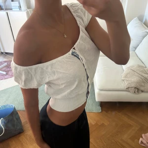 Vit croppad blus med knappar - Superfin vit croppad blus med offshoulder och korta puffärmar. Toppen har knappar framtill, en söt snörning vid halsen och blåvit mönstrad kant. Resår i midjan ger en snygg passform och den är perfekt för varma dagar.