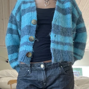 Blå randig kofta - Säljer en mysig blå och grå randig kofta med stora knappar framtill. Koftan är croppad och har långa ärmar. Den är tillverkad i ett mjukt material som ger en skön känsla.