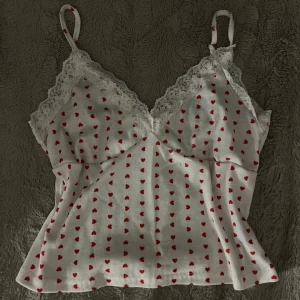 Vit linnetopp med röda hjärtan och spets - Supersöt vit linnetopp med röda hjärtan och tunna axelband. Toppen har v-ringning med vit spetskant och är ribbad i materialet. Perfekt för dig som gillar gulliga detaljer och romantisk stil.