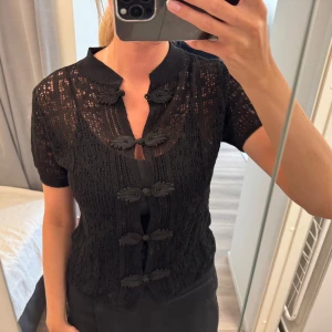 Svart spetsig blus med knappar - Säljer en svart blus i spets med korta ärmar och dekorativa knappar framtill. Kan även användas som en söt kofta med knapparna öppna.