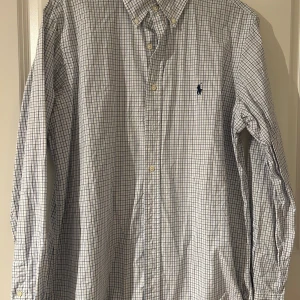 Ralph Lauren skjorta  - Klassisk rutig skjorta från Ralph Lauren – en perfekt blandning av stil och komfort! Denna skjorta är tillverkad i mjukt bomullsmaterial och har den ikoniska Ralph Lauren-loggan på bröstet. Passar lika bra till en avslappnad look med jeans eller uppklädd med chinos för en smart vardagsoutfit. Inga fläckar eller defekter – välvårdad och redo för användning!