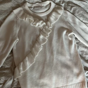 Vit stickad tröja med volang från Vila - Supersöt vit stickad tröja från Vila med lång ärm och snygg volangdetalj som går diagonalt över framsidan. Tröjan har ribbad halsringning och muddar, perfekt för dig som gillar en feminin och trendig look.