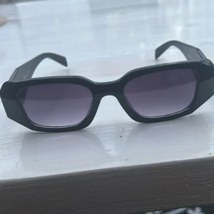 Svarta solglasögon från Prada - Snygga svarta solglasögon från Prada med markerad, kantig båge och mörka glas. Ikonisk logga i guld på sidan och tillverkade i Italien. Perfekt accessoar för en trendig look och att skydda ögonen mot solen.