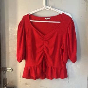 Röd blus  - Superfin röd blus med v-ringning, puffiga halvlånga ärmar och volangkant nertill. Toppen har rynkdetalj framtill och är gjord i ett strukturerat tyg som känns både lätt och luftigt. Perfekt för dig som vill sticka ut med färg och snygga detaljer.