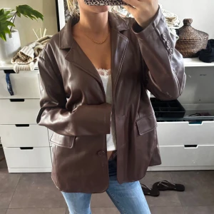 Brun skinnkavaj från Urban Outfitters - Säljer en brun kavaj i skinnimitation från Urban Outfitters. Jackan har klassisk krage, knäppning framtill, fickor med lock och snygga knappar vid ärmslutet. Perfekt oversized fit och riktigt trendig look till jeans eller kjol.