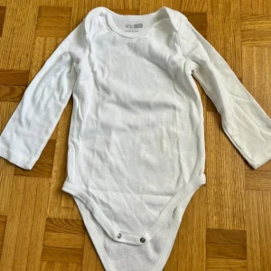 Vit långärmad body från Hello Baby - En basic vit långärmad body från Hello Baby i mjuk bomull. Klassisk rund halsringning och praktiska tryckknappar i grenen för enkel påklädning. Perfekt för lager-på-lager och funkar året runt.