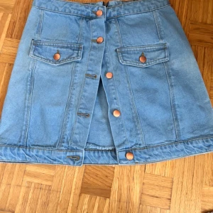 Blå jeanskjol från Missguided - Snygg blå jeanskjol från Missguided med knappar framtill och två fickor med lock och kopparfärgade detaljer. Kjolen är i klassisk denim och har en rak, kort passform som är superenkel att styla till olika looks.