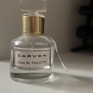 Carven L'Eau de Toilette parfym - Elegant parfymflaska från Carven, L'Eau de Toilette. Flaskan är rund och genomskinlig med en guldig detalj runt halsen och ett stilrent, transparent lock. Etiketten är vit med svart text och ett vitt band hänger från halsen. Perfekt för dig som gillar klassisk och fräsch doft.