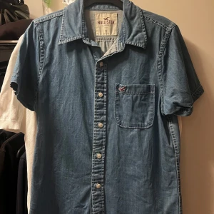 Jeansskjorta från Hollister - Snygg blå skjorta från Hollister i jeanslook med korta ärmar och klassisk krage. Skjortan har knappar framtill och en bröstficka med Hollister-logga. Perfekt för en avslappnad stil och lätt att matcha med andra plagg.