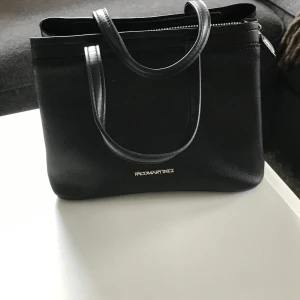 Svart handväska från Paco Martinez - Stilren svart handväska från Paco Martinez i slätt skinn med två handtag och praktisk dragkedja upptill. Väskan har en klassisk fyrkantig form och diskret logga i silver framtill. Perfekt storlek för att få plats med allt du behöver till vardagen.