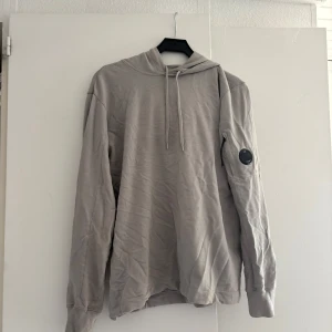 Beige hoodie från C.P. Company - Säljer en stilren beige hoodie från C.P. Company med klassisk huva och snörning. Tröjan har lång ärm och den ikoniska linsen på ena ärmen. Perfekt för dig som gillar enkel och clean streetwear.passar M/L