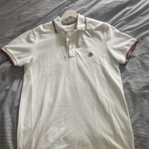 pikétröja Moncler - Vit pikétröja från Moncler med klassisk krage och knäppning framtill. Tröjan har röda och marinblå ränder på krage och ärmslut samt Moncler-logga broderad på bröstet. Tillverkad i bomull och har korta ärmar.