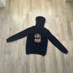 Mörkblå stickad tröja med dinosauriemotiv - Mörkblå hoodie med en stor broderad dinosaurie på bröstet. Tröjan har huva och långa ärmar, samt ribbade muddar vid ärmslut och nederkant. Perfekt för dig som gillar coola och unika motiv. Den är stickad och aldrig använd!💗