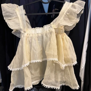 Volangig ljusbeige blus med axelvolanger - Supersöt blus i ljusbeige med breda axelband och stora volanger. Toppen har två lager med volangkant och små bollar längs kanterna. Luftigt, skirt tyg som ger en romantisk vibe. Axelbanden är avtagbara om man vill ha off shoulder💕storlek xs/s. Köptes utomlands. Kan hämtas i Uppsala.