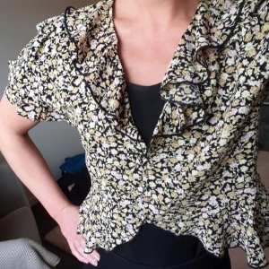 Blommig blus Gina Tricot  - Söt blus från Gina Tricot med ett blommigt mönster i svart, beige och rosa.   Blusen har en volangkrage och volangdetaljer vid ärmarna och nederkanten.  Något kortare med knappslå framtill.  Storlek S/M  Använd vid ett tillfälle, nyskick!  Se gärna mina andra annonser, samfraktar!