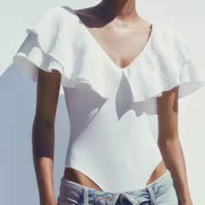 Vit omlott bodysuit med volang - Supersnygg vit bodysuit med bred volang runt axlarna och offshoulder-modell. Säljer då den inte riktigt passa min smak :) aldrig använd! 