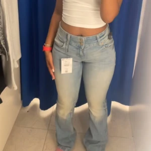 Ljusblå bootcut jeans - Säljer ett par ljusblå bootcut jeans med klassisk femficksdesign och dubbla knappar i midjan. Jeansen har en snygg tvätt och lätt utsvängda ben som ger en retrovibe. Perfekta att styla med crop top eller hoodie.