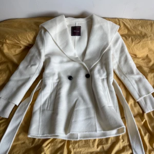 Vit kappa från Alessa - Snygg vit SHAWL COLLAR CASHMERE COAT  från Alessa med bred krage, knytskärp i midjan och två knappar framtill. Jackan har långa ärmar med ribbade muddar och raka fickor. Perfekt för dig som vill ha en stilren och mysig look under kyliga dagar.