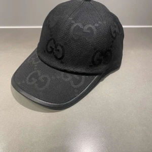 Svart keps från Gucci med GG-mönster - Snygg svart keps från Gucci med klassiskt GG-mönster ton-i-ton över hela kepsen. Justerbar rem baktill och böjd skärm. Tillverkad i slitstark bomull och polyester, perfekt för dig som vill ha en stilren och lyxig accessoar.