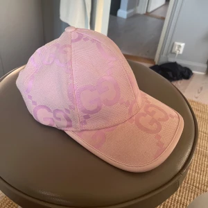 Rosa Gucci keps - Exklusiv rosa keps från Gucci med ikoniskt GG-mönster i ljuslila. Kepsen är tillverkad i en lyxig blandning av 90% merinoull och 10% kashmir, med justerbar rem bak i ljusrosa läder. Perfekt accessoar för dig som vill sticka ut med stil. 