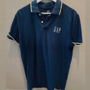 Mörkblå GAP pikétröja med gul kant - Snygg mörkblå pikétröja från GAP med korta ärmar och gul kant på krage och ärmslut. Klassisk polo med knappar framtill och broderad GAP-logga på bröstet. Perfekt för en clean och sportig look.