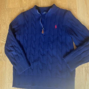 Marinblå kabelstickad tröja Polo Ralph Lauren - Kabelstickad marinblå tröja från Polo Ralph Lauren med halv dragkedja och röd broderad logga på bröstet. Tröjan har ribbade muddar och är tillverkad i 100% bomull. Perfekt för lager-på-lager och snygg till jeans. Den har krympt lite i tvätten så den passar mer strl S