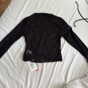 Svart spetsblus från Cider - Säljer en svart långärmad blus från Cider i transparent spets med blommigt mönster. Toppen är figurnära och har en lätt croppad passform. Perfekt för dig som vill ha en edgy och trendig look med snygga detaljer.