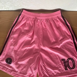 Inter Miami Messi Shorts - Rosa fotbollsshorts från Inter Miami med svarta detaljer och klubbmärke på ena benet. Snygga svarta ränder längs sidorna och tryckt nummer 10. Tillverkade i lätt och ventilerande polyester, perfekta för fotbollsträning eller match.   Storlek XL. Kan hämtas i Vallentuna.