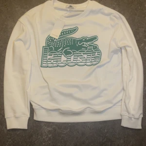 Vit sweatshirt från Lacoste med logga - Vit sweatshirt från Lacoste med stor grön krokodil och logga framtill. Klassisk rund halsringning och långa ärmar. Mjuk bomullskänsla och ribbade muddar vid ärmslut och nederkant. Perfekt för en clean och sportig look.
