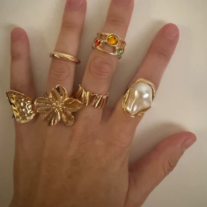 Set med 6 chunky guldiga ringar - Säljer ett set med 6 statementringar i guldton. En har stor blomform, en med pärlliknande detalj, en med färgade stenar, samt flera med coola, organiska former. Perfekt för dig som gillar att mixa och matcha smycken och sticka ut.
