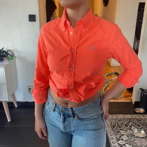 Orange skjorta från Ralph Lauren - Snygg orange skjorta från Ralph Lauren med klassisk button down-krage och blå broderad logga på bröstet. Skjortan är långärmad, har knappar hela vägen och är i bomull. Passar både öppen och knuten för en chill vibe. Nya modellen❤️