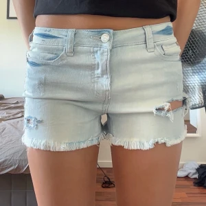 Ljusblå jeansshorts med slitningar - Snygga ljusblå jeansshorts med fransig kant och slitna detaljer framtill. Klassisk femficksmodell med knapp och dragkedja. Perfekta för varma dagar och ger en avslappnad vibe. Aldrig använda och i nyskick!