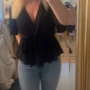 Svart omlottblus med knytband - Snygg svart omlottblus med v-ringning och knytband i midjan. Blusen har korta ärmar och volangkant nedtill som ger en feminin siluett. Perfekt att styla med jeans eller kjol för en trendig look.