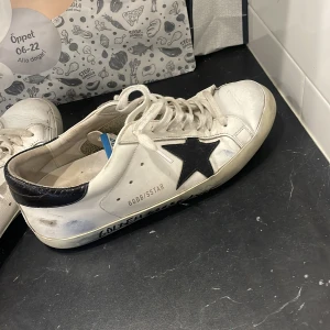 Golden Goose 42 - Golden Goose sneakers i vitt skinn med ikonisk svart stjärna på sidan och svart häl. Klassisk låg modell med snörning och platt sula. Snyggt slitna detaljer och handskriven text på sulan ger en cool, urban vibe.