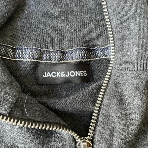 Grå half zip tröja Jack & Jones M - Mörkgrå stickad tröja från Jack & Jones i storlek M. Tröjan har hög krage och en snygg half zip-dragkedja framtill. Materialet är mjukt och ribbstickat vid ärmslut och nederkant, perfekt för lager på lager under kyliga dagar.