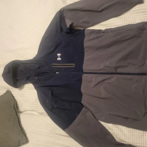 Jacka Under armour  - Vindjacka från Under Armour. Jackan har huva med dragkedja. Se mått på bilder. Dragkedjan skadad men funkar fortfarande.  Därav priset. Se gärna mina andra annonser och skapa bundels.