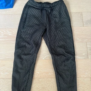 Svarta Nike mjukisbyxor med mönster - Nike mjukisbyxor i svart med små rutigt mönster och diskret logga på benet. Byxorna har resår och snörning i midjan, dragkedjeficka och muddar vid benslut. Perfekta för chill eller träning, riktigt sköna och snygga.