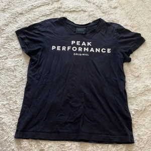 Svart Peak Performance t-shirt - Svart t-shirt från Peak Performance med vit logga tryckt på bröstet. Klassisk rund halsringning och korta ärmar. Tillverkad i mjuk bomull som är skön att ha på sig hela dagen. Perfekt för en sportig och avslappnad stil.