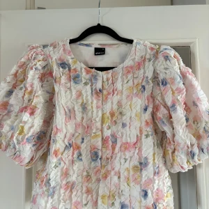 Blommig volangblus  - Supersöt blus med blommigt mönster i pastellfärger från Gina Tricot. Puffiga korta ärmar och volangstruktur över hela toppen. Basen är vit med inslag av rosa, blått och gult.