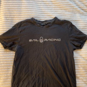 Svart t-shirt från Sail Racing - Svart t-shirt från Sail Racing med tryckt logga i vitt över bröstet. Klassisk rund halsringning och korta ärmar. Skön och enkel stil som funkar till det mesta. Bara att dm:a vid frågor eller funderingar💕