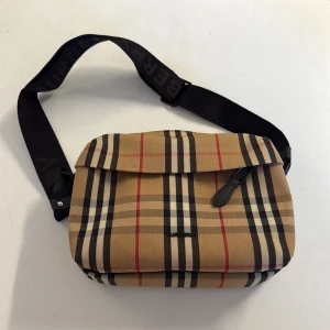 Rutig axelremsväska från Burberry - Säljer en klassisk axelremsväska från Burberry i beige, svart, röd och vit rutig design. Väskan har justerbar svart axelrem med logga, dragkedja upptill och innerfack med kortplatser i svart skinn. Perfekt för dig som gillar stilrena accessoarer med ikoniskt mönster.