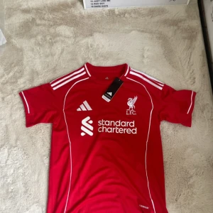 Liverpool FC hemmatröja 25/26 - Röd Liverpool FC fotbollströja från Adidas i slim fit. Klassisk design med vita detaljer, klubbmärke och sponsortryck på bröstet. Tröjan har korta ärmar och är tillverkad i lätt och ventilerande polyester, perfekt för match eller träning.
