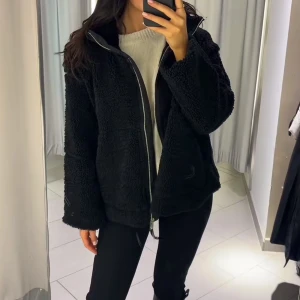 Svart teddyjacka med dragkedja - Mysig svart teddyjacka med hel dragkedja framtill och hög krage. Jackan har en loose passform och är tillverkad i fluffigt teddy-material som håller dig varm. Perfekt för kyliga dagar och enkel att styla med dina favoritjeans eller leggings. Extrem lik den från hm nästan iddentiska